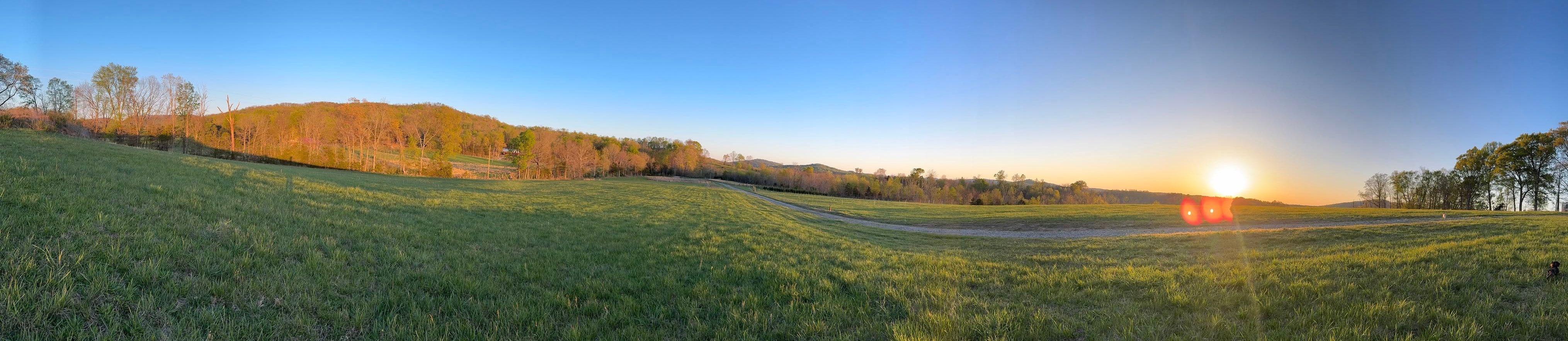 A wide view across the farm pasture at sunset, with a gravel drive curving through the grass and wooded hills behind.